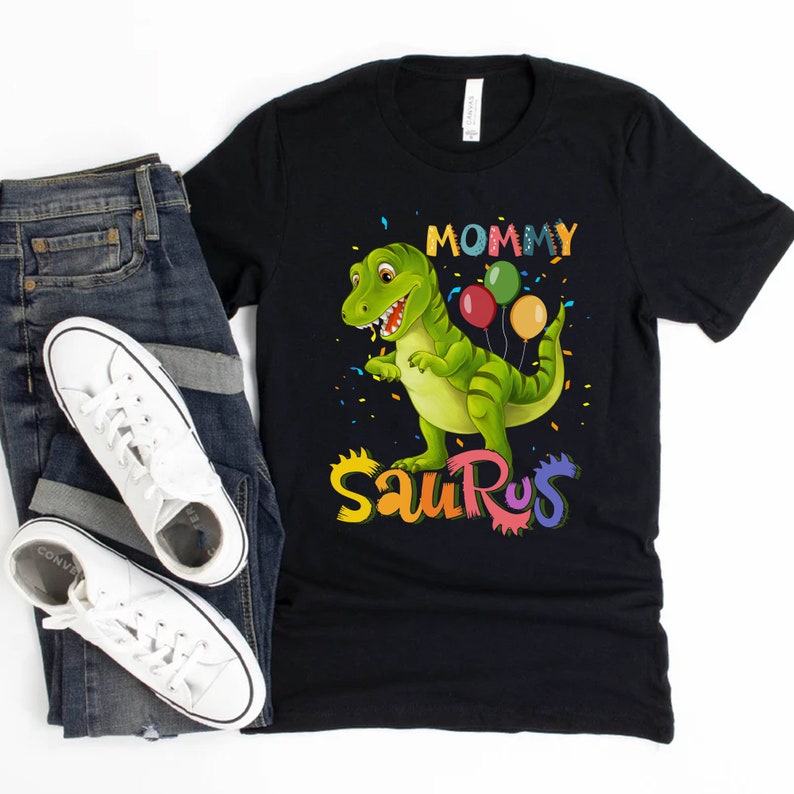 May include: Black t-shirt featuring a green dinosaur holding colourful balloons. The text "Mommy Saurus" is written in rainbow colours.