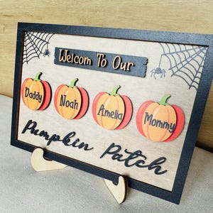 Pode incluir: Uma placa retangular de madeira com moldura preta apresentando o texto Welcom To Our Pumpkin Patch. Quatro abóboras laranja são exibidas, cada uma rotulada com os nomes Daddy, Noah, Amelia e Mommy. O fundo inclui ilustrações de teias de aranha pretas.