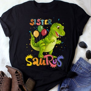 May include: Black t-shirt featuring a green dinosaur holding balloons and the text "Sister Saurus" in rainbow colours.