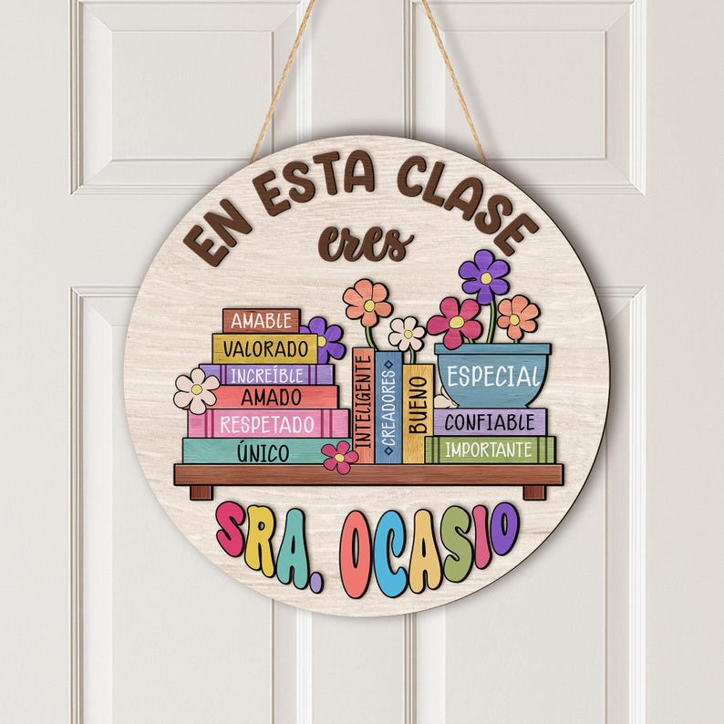 May include: A round wooden sign with the words "EN ESTA CLASE eres" in brown lettering. The sign features a shelf with books and flowers, and the name "SRA. OCASIO" in colorful letters.