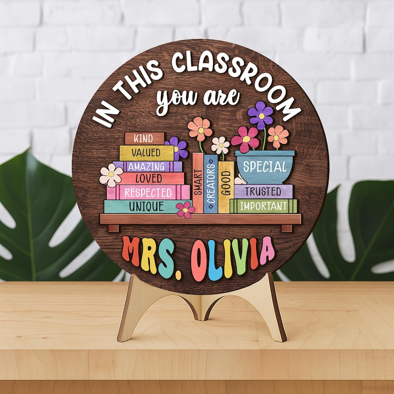 Puede incluir: Un letrero de aula de madera con el texto "IN THIS CLASSROOM YOU ARE" y "MRS. OLIVIA". El letrero presenta una estanter&iacute;a con libros y flores de colores. Tambi&eacute;n son visibles las palabras "KIND, VALUED, AMAZING, LOVED, RESPECTED, UNIQUE, SMART, CREATORS, GOOD, SPECIAL, TRUSTED, IMPORTANT".