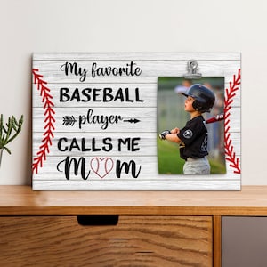 May include: White wood photo frame with a baseball theme. The frame has a red stitched baseball seam design on the sides. The text reads "My favorite baseball player calls me mom" with a baseball and heart graphic. A photo of a young boy holding a baseball bat is in the frame.