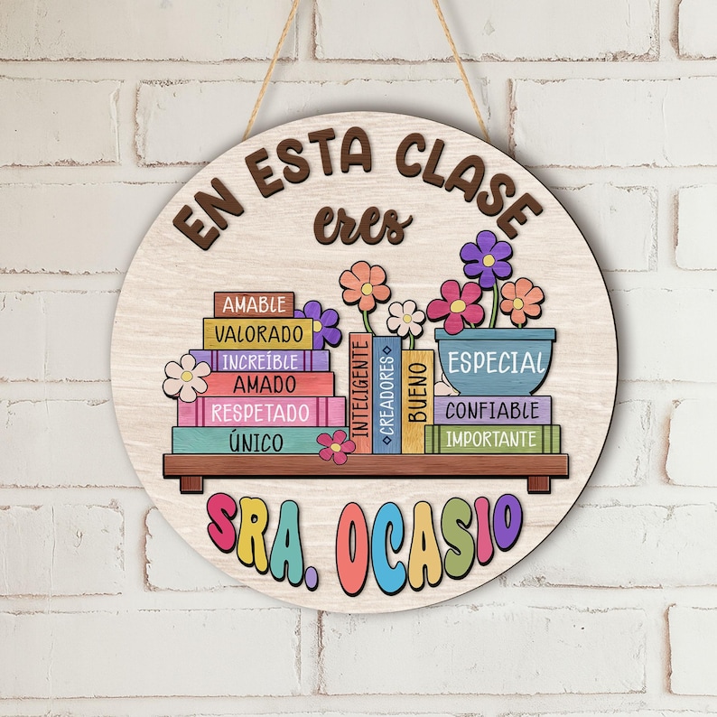 May include: A round wooden sign with the words "EN ESTA CLASE ERES" in brown lettering. Below, colorful books with positive words like "AMABLE" and "ESPECIAL" are displayed. The sign is decorated with flowers and the name "SRA. OCASIO".