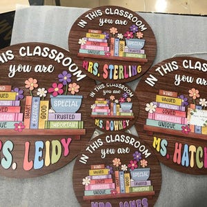 Puede incluir: Carteles de aula de madera con la frase "En esta clase eres" y el nombre de un profesor. Cada cartel presenta una estanter&iacute;a con libros coloridos, flores y palabras positivas como "Amable" y "Amado".