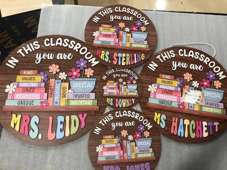 Puede incluir: Carteles de aula de madera con la frase "En esta clase eres" y el nombre de un profesor. Cada cartel presenta una estanter&iacute;a con libros coloridos, flores y palabras positivas como "Amable" y "Amado".