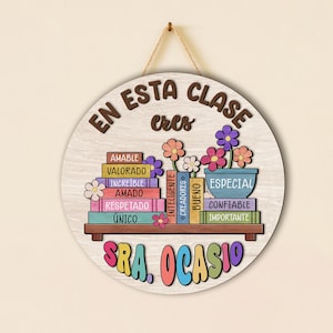 May include: Round wooden sign with the text "EN ESTA CLASE eres" in brown lettering. The sign features a shelf with colorful books and flowers, with the name "SRA. OCASIO" in bright colors at the bottom. The sign is hanging by a natural twine.