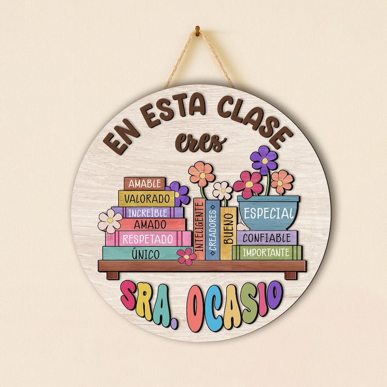 May include: Round wooden sign with the text "EN ESTA CLASE eres" in brown lettering. The sign features a shelf with colorful books and flowers, with the name "SRA. OCASIO" in bright colors at the bottom. The sign is hanging by a natural twine.