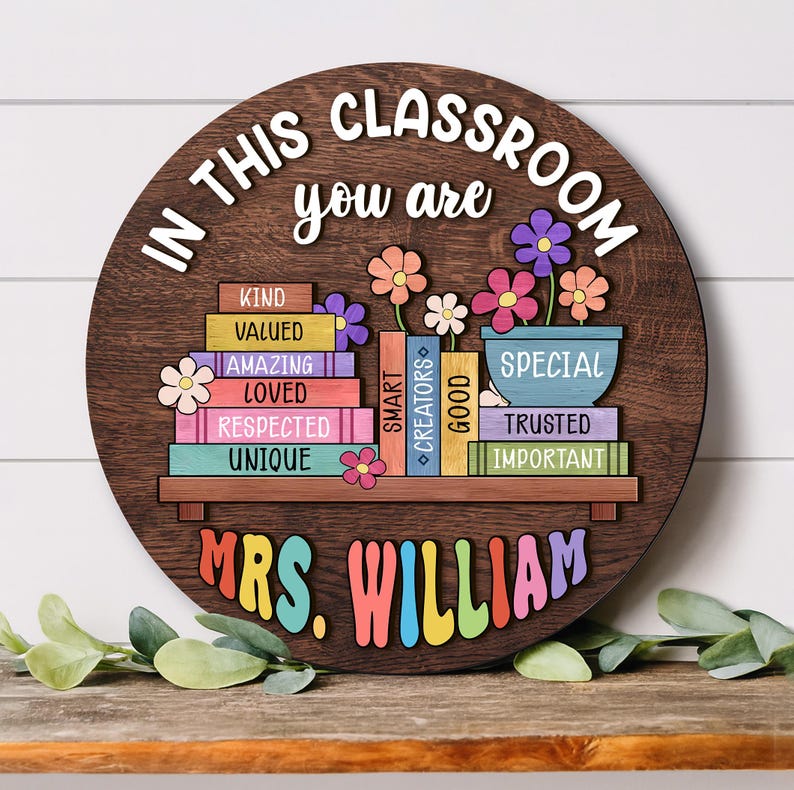 Puede incluir: Un letrero redondo de madera con el texto "IN THIS CLASSROOM you are" y "MRS. WILLIAM". El dise&ntilde;o presenta una estanter&iacute;a con libros coloridos y flores. Los libros muestran palabras como "KIND", "VALUED" y "SPECIAL".