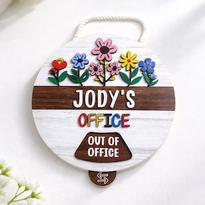 Puede incluir: Un letrero redondo de madera con fondo blanco y una franja marrón. Presenta flores coloridas, el texto "JODY'S OFFICE" y un indicador reversible "OUT OF OFFICE". Una cuerda está unida para colgarlo.