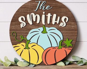Puede incluir: Un letrero circular de madera con acabado de veta de madera marrón oscuro que presenta el texto The Smiths en azul claro y crema. Debajo del texto hay tres calabazas de madera en capas en azul claro, amarillo y naranja con tallos verdes.