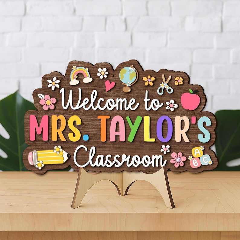 Puede incluir: Un letrero de aula de madera con el texto "Welcome to MRS. TAYLOR'S Classroom". El letrero es marr&oacute;n con acentos coloridos, incluyendo un arco&iacute;ris, flores y un l&aacute;piz. Se apoya en un caballete de madera.