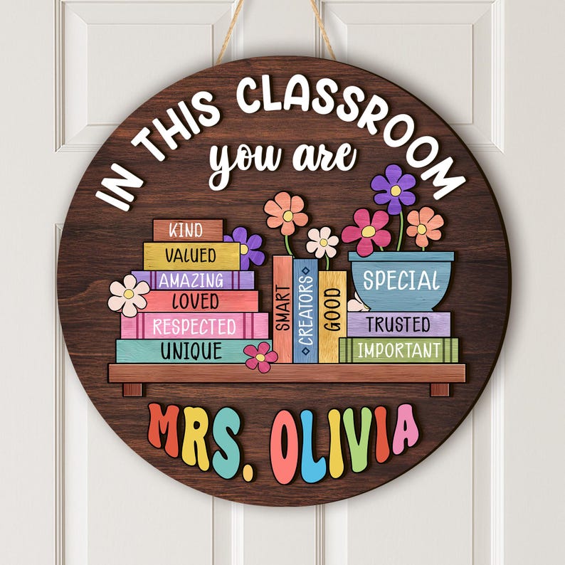 Puede incluir: Un letrero de aula redondo de madera con el texto "IN THIS CLASSROOM you are" en blanco. El letrero presenta libros coloridos, flores y el nombre "MRS. OLIVIA" en colores del arco&iacute;ris. Cuelga de una puerta blanca.