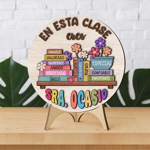 May include: A wooden sign with the text "EN ESTA CLASE eres" and "SRA. OCASIO". The sign features a shelf with colorful books and flowers. The books have words like "AMABLE" and "ESPECIAL". The sign is on a wooden stand.
