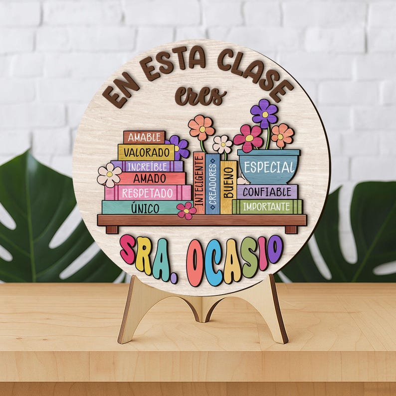 May include: A wooden sign with the text "EN ESTA CLASE eres" and "SRA. OCASIO". The sign features a shelf with colorful books and flowers. The books have words like "AMABLE" and "ESPECIAL". The sign is on a wooden stand.