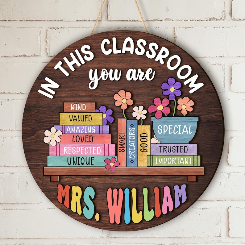 Puede incluir: Un letrero redondo de madera con el texto "IN THIS CLASSROOM you are" y "MRS. WILLIAM". El letrero presenta un dise&ntilde;o de estanter&iacute;a con libros y flores de colores. Los libros tienen palabras como "KIND", "VALUED" y "SPECIAL".