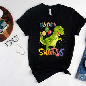 May include: Black t-shirt featuring a green dinosaur holding colourful balloons and the text "Daddy Saurus" in rainbow colours.