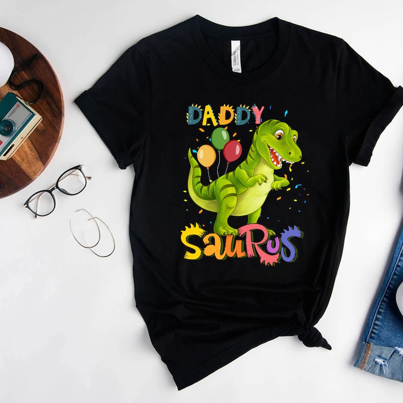 May include: Black t-shirt featuring a green dinosaur holding colourful balloons and the text "Daddy Saurus" in rainbow colours.