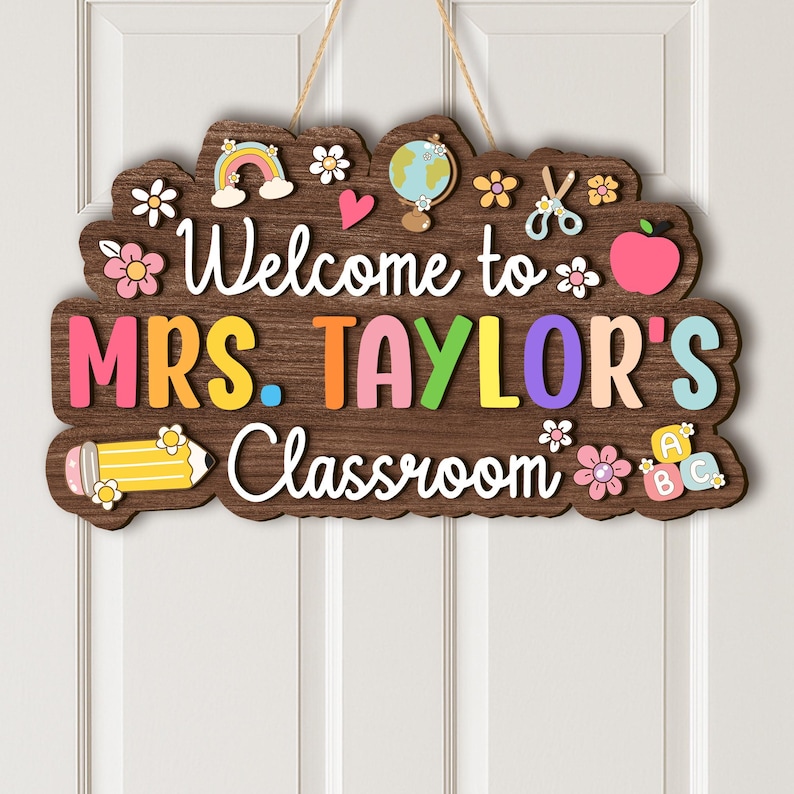Puede incluir: Un letrero de aula de madera con el texto "Welcome to MRS. TAYLOR'S Classroom" en blanco, con detalles coloridos. El letrero incluye un arco&iacute;ris, un globo terr&aacute;queo, una manzana y un l&aacute;piz. Cuelga de una cuerda natural.