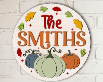 Puede incluir: Un letrero redondo de madera con acabado de veta blanca, que presenta el texto The SMITHS en rojo y naranja. El diseño incluye tres calabazas en azul, verde claro y naranja, rodeadas de pequeñas hojas coloridas y estrellas rojas.
