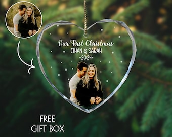 May include: A heart-shaped clear glass Christmas ornament with a photo of a couple inside. The ornament has the text "Our First Christmas ETHAN & SARAH -2024-" and snowflake details. A gold string is attached for hanging.