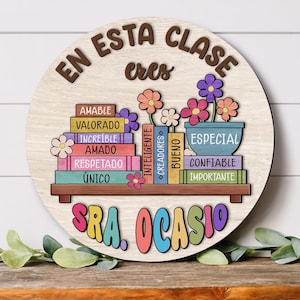 Op de afbeelding: Een ronde houten plaat met de woorden "EN ESTA CLASE eres" in bruine letters. Daaronder kleurrijke boeken met positieve woorden en een bloempot met bloemen. De naam "SRA. OCASIO" staat in kleurrijke letters onderaan.