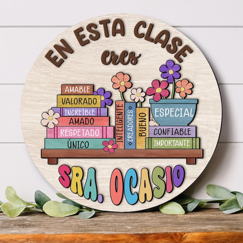 May include: A round wooden sign with the words "EN ESTA CLASE eres" in brown letters. Below, colorful books with positive words and a flower pot with flowers. The name "SRA. OCASIO" is written in colorful letters at the bottom.