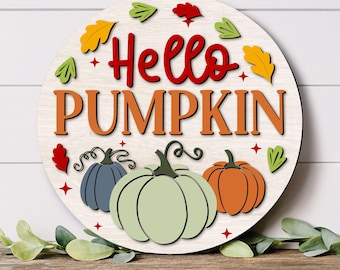 Puede incluir: Un letrero redondo de madera con fondo de veta de madera blanca que presenta el texto rojo Hello y el texto naranja PUMPKIN. El diseño incluye tres calabazas en azul, verde claro y naranja, acentuadas por pequeñas estrellas rojas y hojas coloridas.