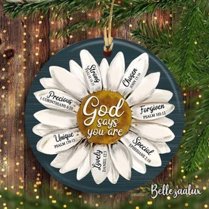 May include: A round, navy blue ornament featuring a white daisy design with the words "God says you are" in the center. The daisy petals have words like "Strong", "Chosen", and "Precious". A gold string is attached for hanging.