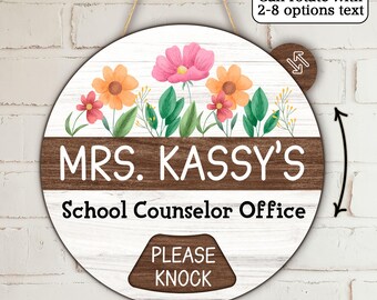 May include: Round wooden sign with floral accents and the text "MRS. KASSY'S School Counselor Office." Features a rotating section with text options and a "PLEASE KNOCK" sign. The sign is suspended by a natural twine.