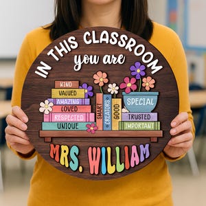 Puede incluir: Un letrero redondo de madera con el texto "IN THIS CLASSROOM you are" y "MRS. WILLIAM". El letrero presenta libros apilados coloridos con palabras positivas y acentos florales. Un regalo de agradecimiento para profesores.