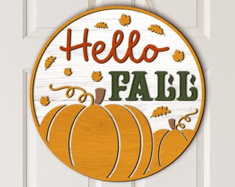 Puede incluir: Un letrero de puerta redondo de madera con fondo blanco que imita tablones. El texto "Hello FALL" está escrito en letras naranjas y verde oscuro. Dos calabazas naranjas con tallos marrones y pequeñas hojas dispersas decoran la parte inferior.