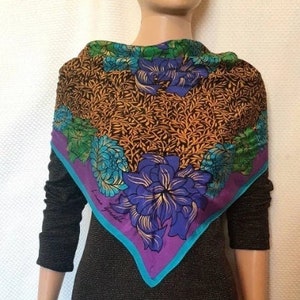 Puede incluir: Un pañuelo cuadrado de seda con un diseño floral. El pañuelo presenta un fondo marrón con patrones de hojas doradas y grandes flores azules y turquesas. El borde es morado con un ribete turquesa. El pañuelo está drapeado alrededor de un maniquí.