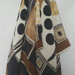 May include: A patterned scarf with a mix of geometric and abstract designs. The scarf features large black polka dots, brown squares, and cream and tan sections. The scarf is hanging against a white background.