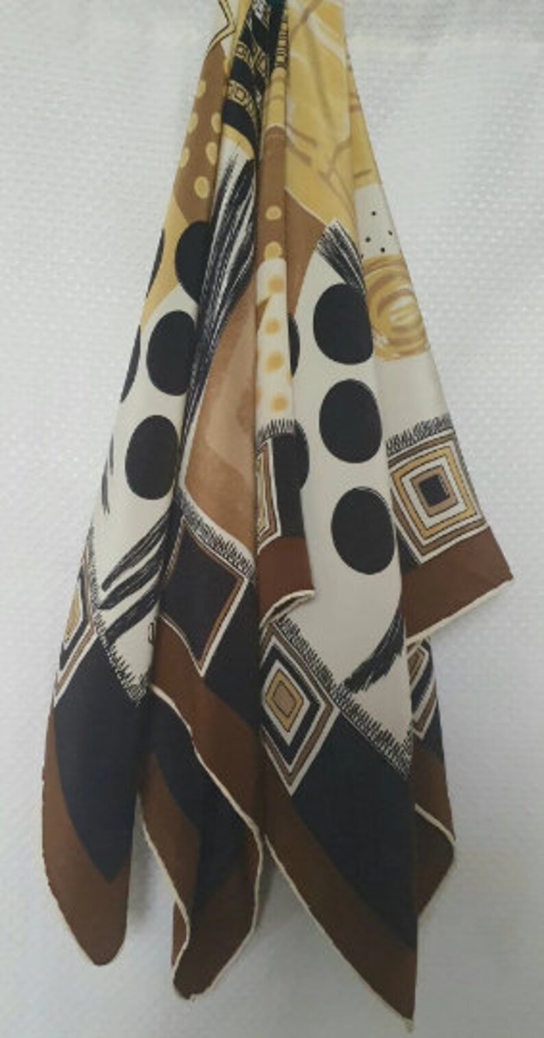 May include: A patterned scarf with a mix of geometric and abstract designs. The scarf features large black polka dots, brown squares, and cream and tan sections. The scarf is hanging against a white background.