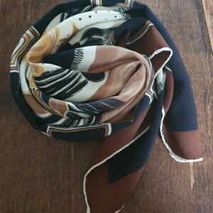 May include: A folded silk scarf with a geometric and abstract design. The scarf features a color palette of navy blue, brown, cream, and gold. The edges are finished with a white trim.