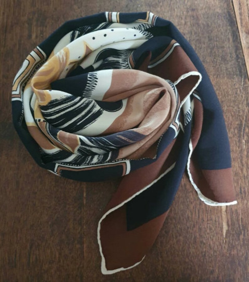 May include: A folded silk scarf with a geometric and abstract design. The scarf features a color palette of navy blue, brown, cream, and gold. The edges are finished with a white trim.