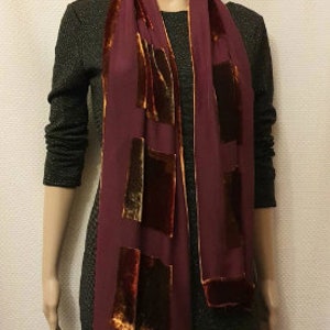 May include: A burgundy scarf with rectangular velvet accents and trim. The scarf is displayed on a mannequin, highlighting its design and texture. The velvet patches add a touch of luxury to the accessory.