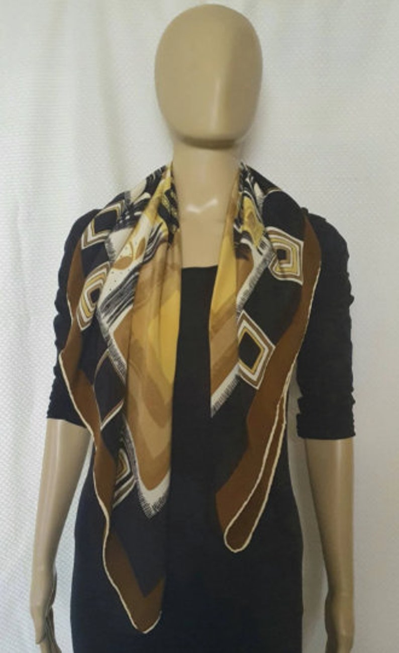 May include: A square scarf with a geometric pattern in black, brown, white, and yellow. The scarf is draped around a mannequin's neck, showcasing its design. The scarf's edges are trimmed with a brown border.