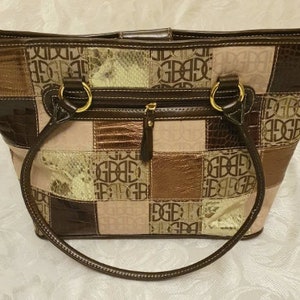 Op de afbeelding: Een patchwork tote bag met bruine, beige en bronzen vierkanten. De tas heeft een ritssluiting, goudkleurige ringen en handvatten. Het patroon "GBGB" herhaalt zich over het hele oppervlak.