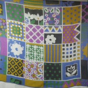 May include: A square scarf with a patchwork design featuring various geometric and floral patterns. The scarf has a blue border and a mix of colors including green, purple, yellow, and white. The design includes polka dots, stripes, and flower motifs.