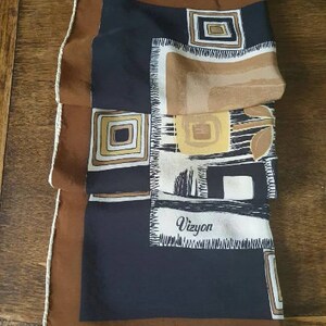May include: A folded silk scarf with a brown border and a white stitched edge. The scarf features a geometric design with squares and rectangles in shades of brown, tan, black, and white. The word "Vizyon" is printed on the scarf.