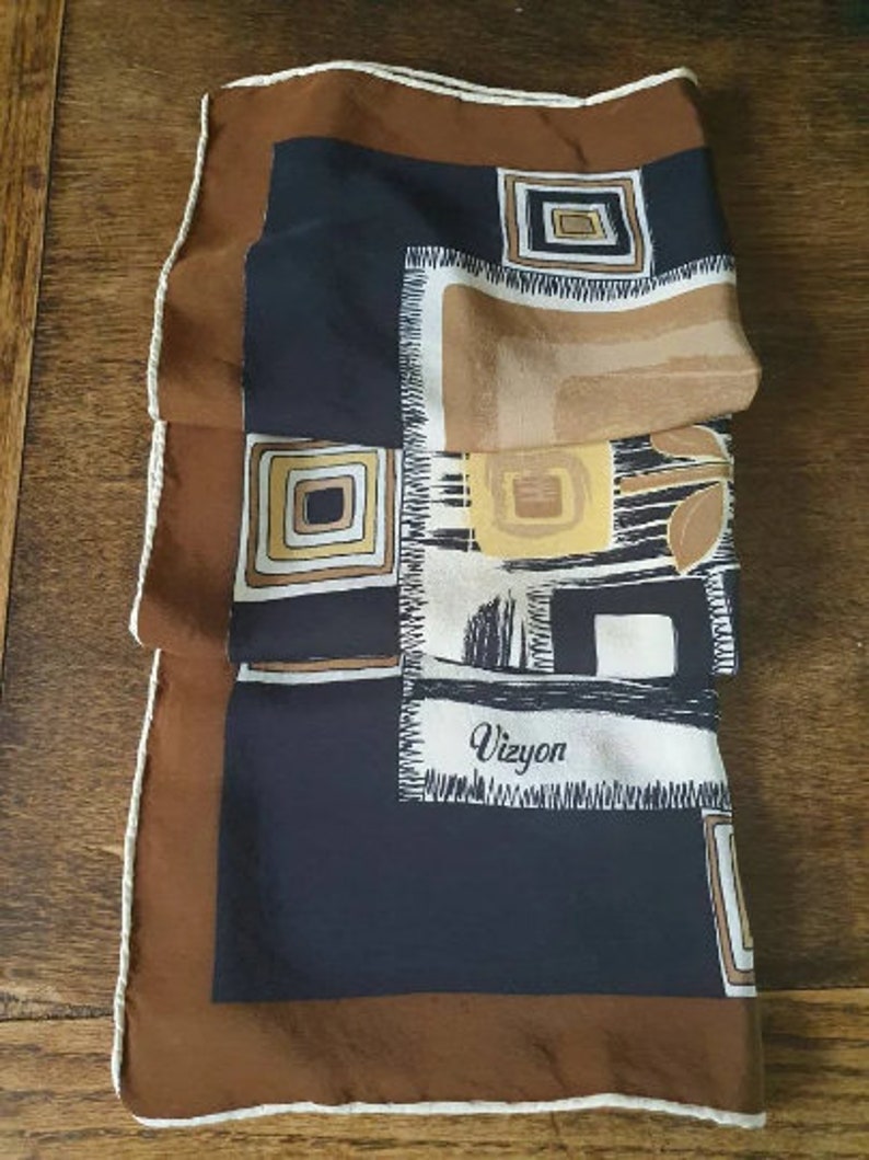 May include: A folded silk scarf with a brown border and a white stitched edge. The scarf features a geometric design with squares and rectangles in shades of brown, tan, black, and white. The word "Vizyon" is printed on the scarf.