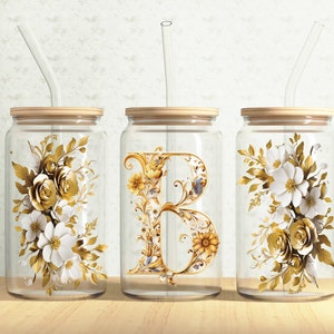 Könnte beinhalten: Drei gläserne Becher mit Holzdeckeln und klaren Strohhalmen. Jeder Becher hat ein anderes Design: ein Blumenmuster mit weißen und goldenen Blumen, ein goldener floralen Buchstabe B und ein Blumenmuster mit weißen und goldenen Blumen.
