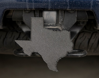 May include: A black metal Texas state outline hitch cover. The hitch cover is attached to a black vehicle.