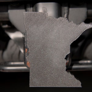 May include: A gray 3D printed outline of the state of Minnesota attached to a black metal hitch receiver.