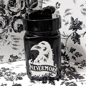 May include: A black glass bottle with a black screw top lid. The bottle has a white raven graphic with the words 'NEVERMORE' printed below the raven.