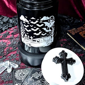 May include: Black jar with a white graphic of bats, clouds, and a hearse. A white lid with a black cross sits next to the jar. Tarot cards and crystals are also visible.