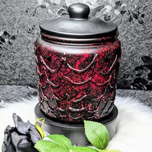 Pode incluir: Um frasco decorativo com tampa, apresentando um design salpicado em vermelho e preto. O frasco está sobre uma base preta, com uma rosa preta e folhas verdes por perto. O fundo é um padrão floral em tons de cinza.