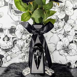 Op de afbeelding: Een zwarte decoratieve vaas met een raven schedel ontwerp, met zwarte rozen en groene bladeren, vastgebonden met een zwart lint. Drie tarotkaarten liggen voor de vaas, met een schedel- en bloemenachtergrond.
