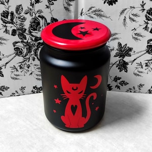 May include: Black glass jar with a red lid and a red silhouette of a cat with a crescent moon and stars. The jar is decorated with a red crescent moon and stars on the lid.